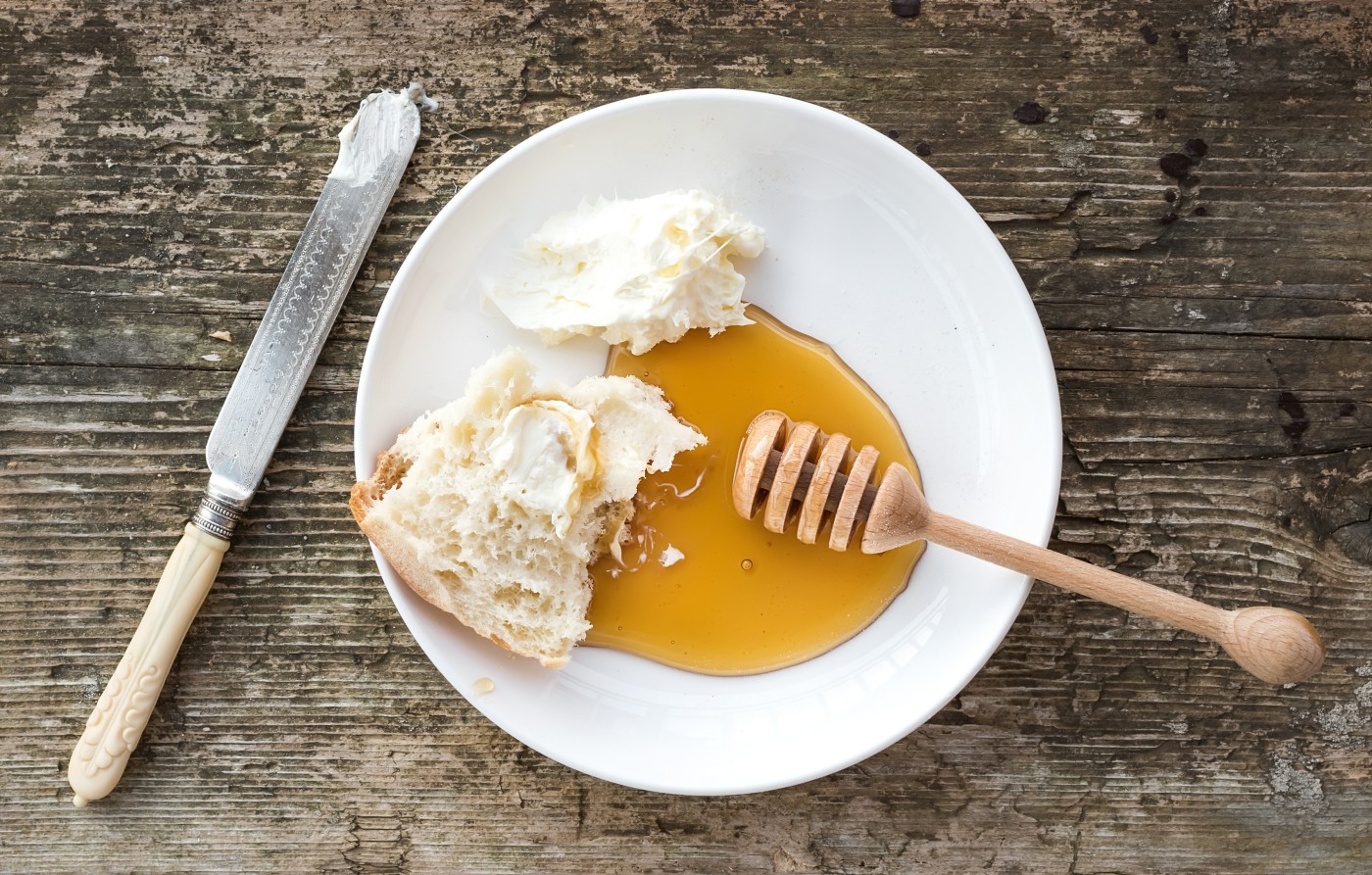 Tradirional Balkan breakfast set: kaymak or butter cream, flatbread and honey on a white ceramic plate with a knife over a rough wood background. Top view